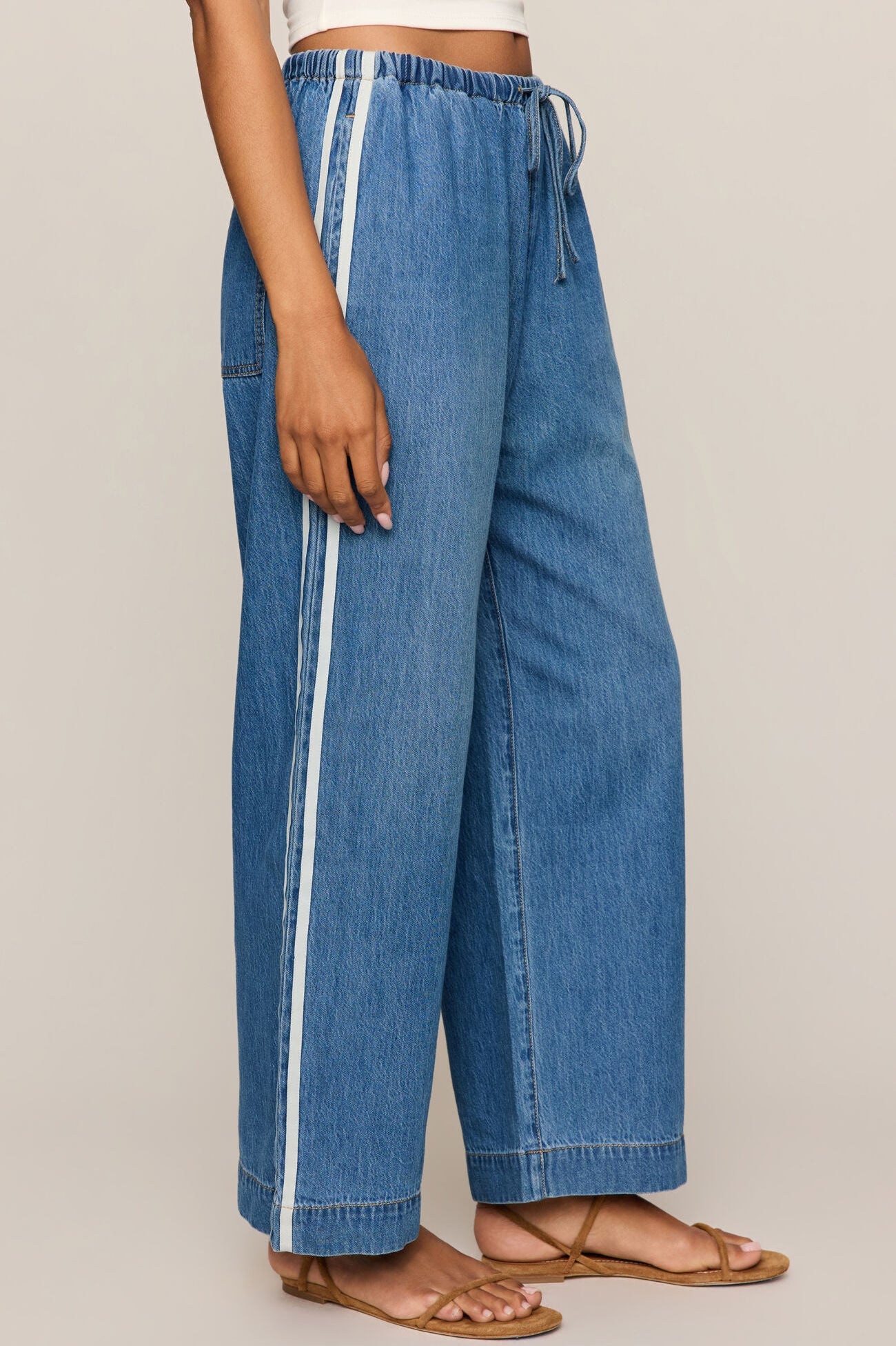 Side view of woman wearing a white tank top and sidelines denim track pant on a beige background