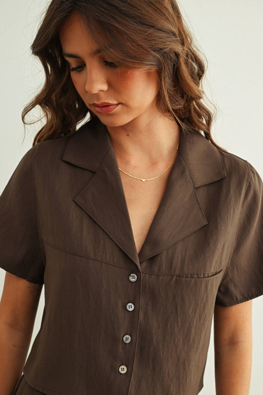 A woman wearing a brown short-sleeve button-front shirt with notched lapels, shown from the chest up against a plain light background.