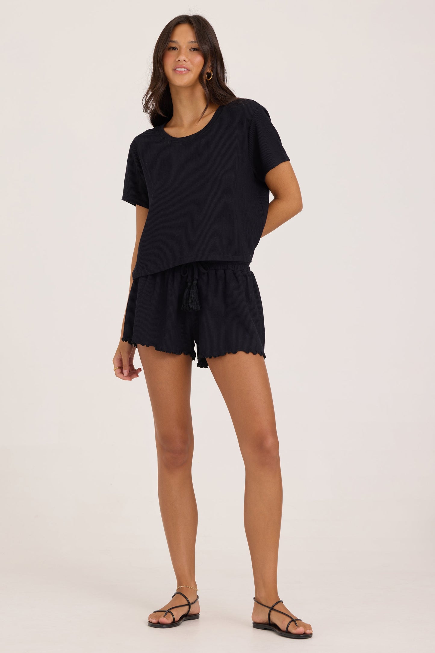 A woman modeling a black short-sleeve t-shirt and matching black smock waist drawstring shorts with scalloped hems, paired with black strappy sandals, against a plain light background.
