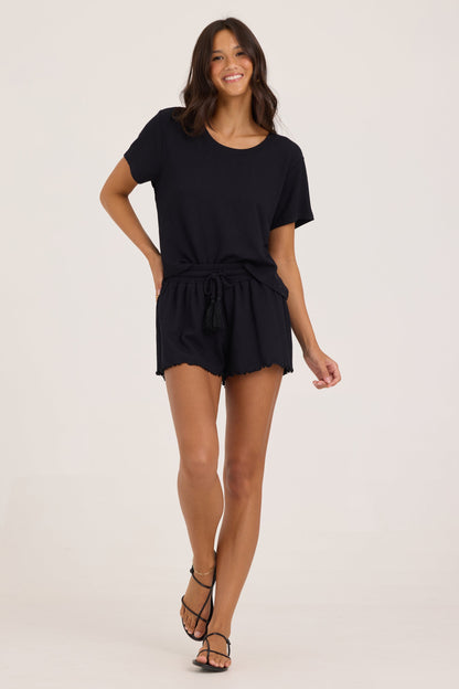 A woman modeling a black short-sleeve shirt with black smock waist drawstring shorts, photographed against a plain light beige studio background.