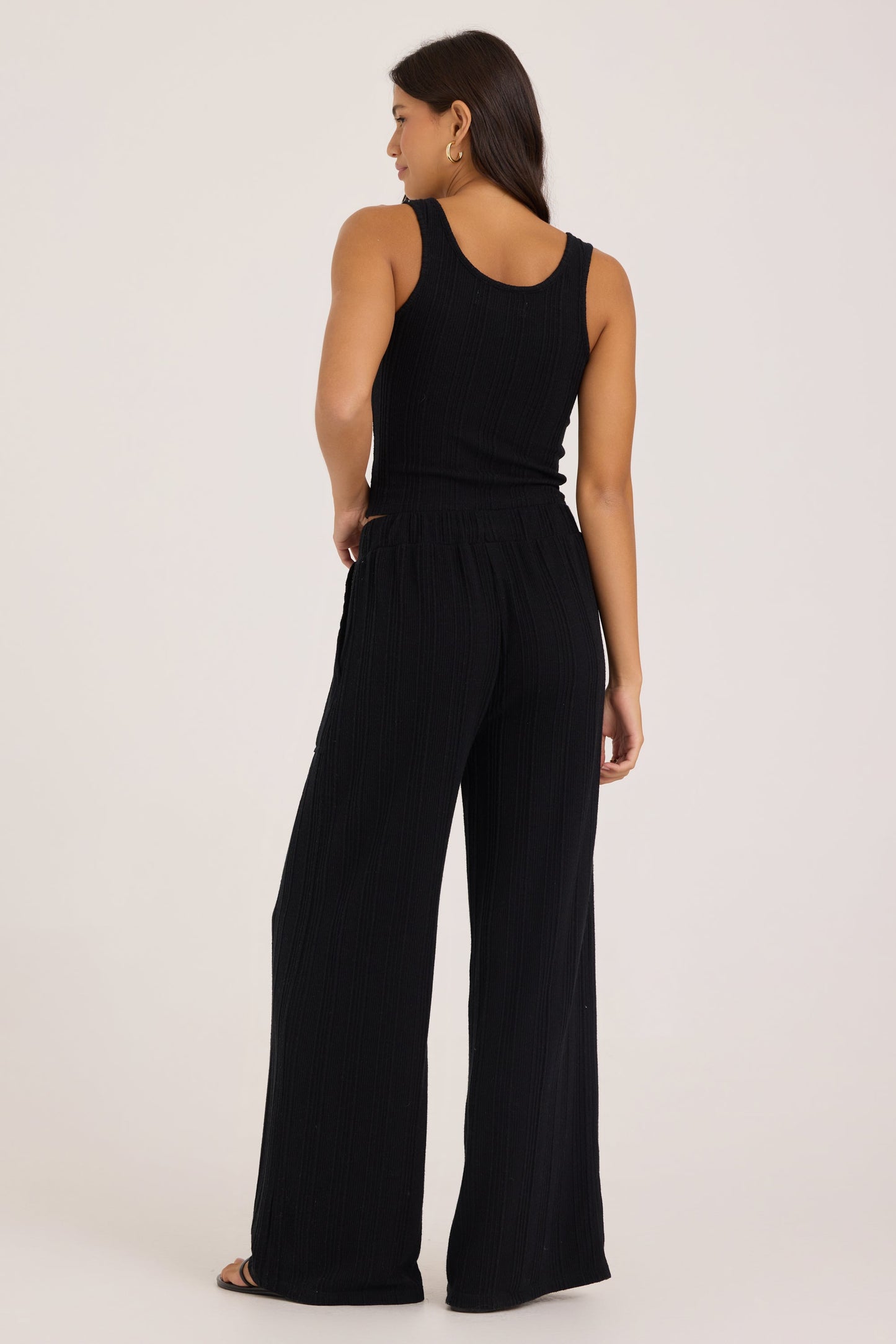 A woman shown from the back wearing a black sleeveless tank with wide-leg pants, photographed against a plain light beige studio background.