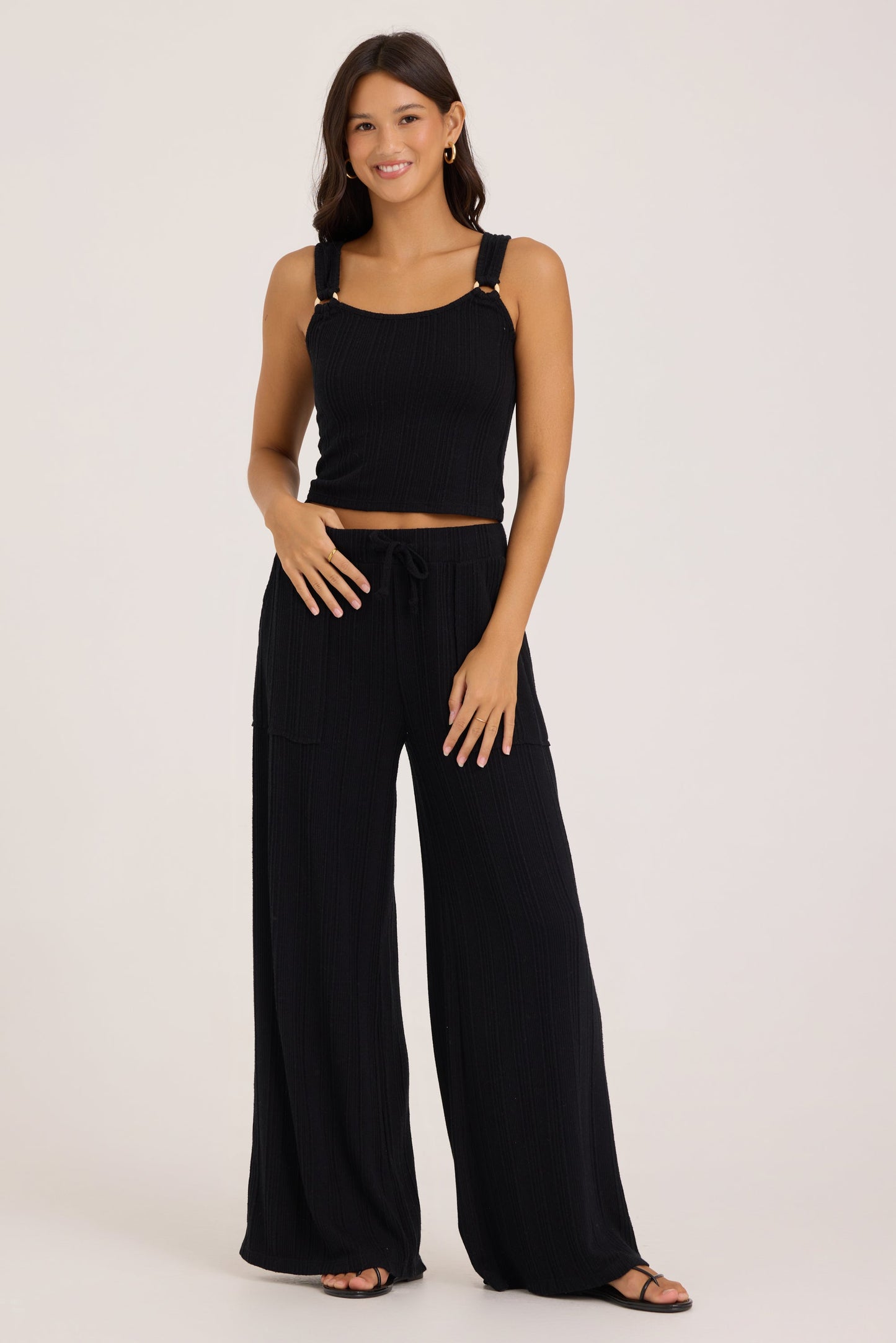 A woman modeling a black ribbed knit two-piece set consisting of a cropped o-ring tank top with gold-tone shoulder hardware and matching wide-leg drawstring pants, photographed against a plain light studio background.