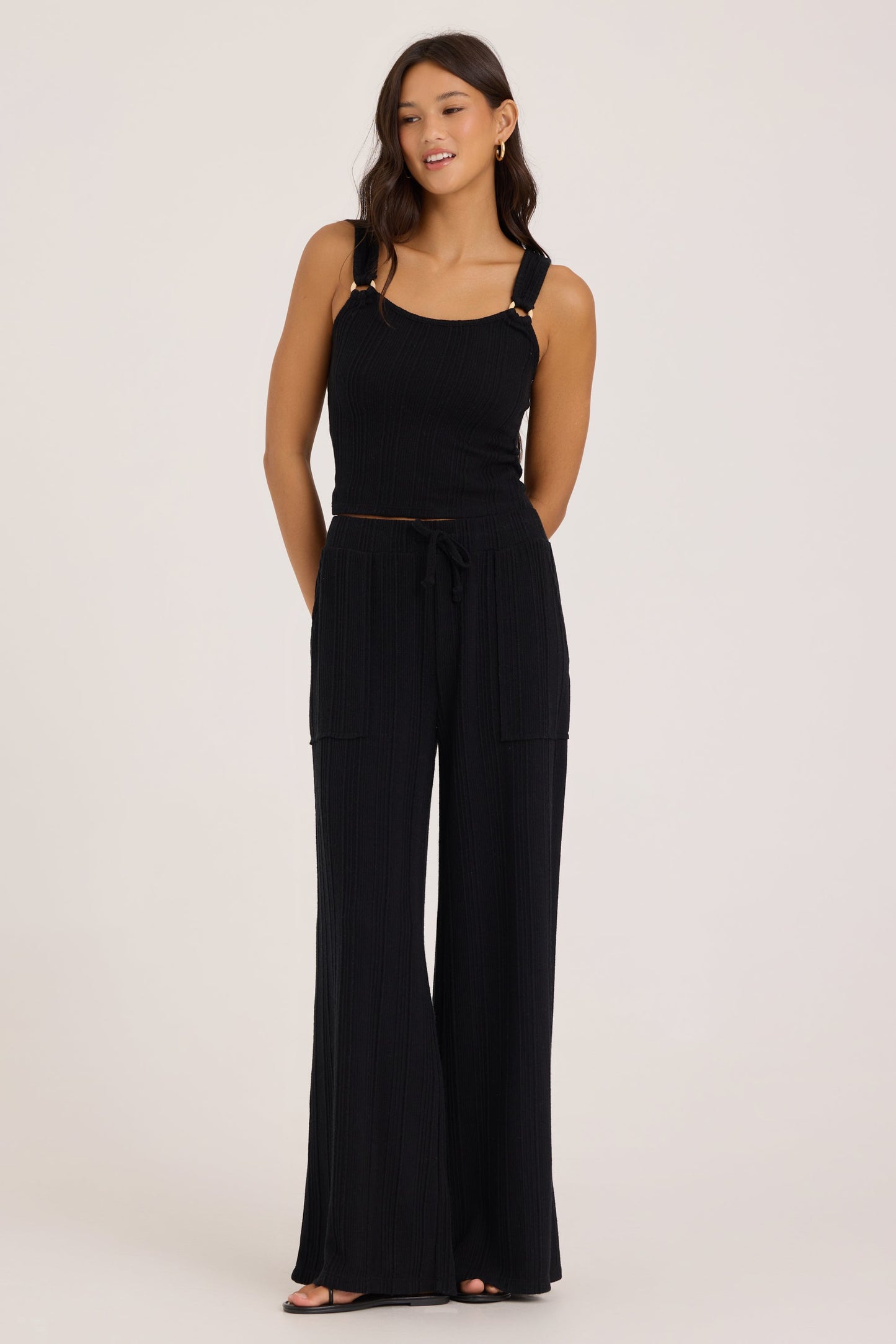 A woman modeling a black sleeveless jumpsuit with wide-leg pants, a tie waist, and gold-tone o-ring hardware on the shoulder straps, photographed against a plain light gray studio background.