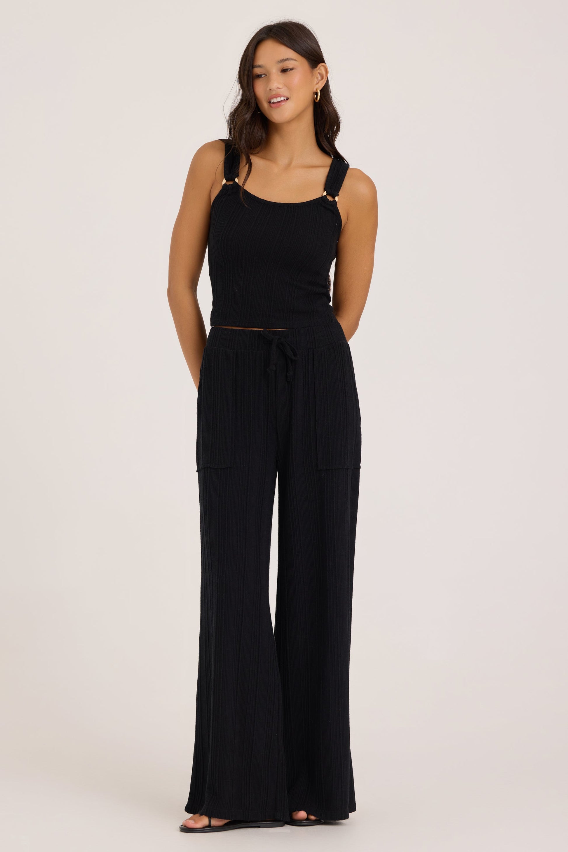 A woman modeling a black sleeveless jumpsuit with wide-leg pants, a tie waist, and gold-tone o-ring hardware on the shoulder straps, photographed against a plain light gray studio background.