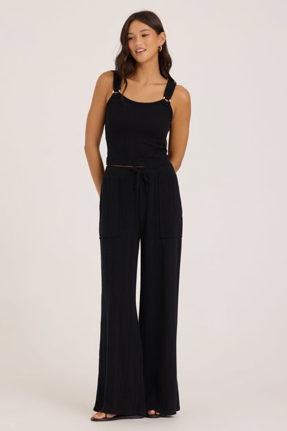 A woman modeling a black sleeveless jumpsuit with wide-leg pants, a tie waist, and gold-tone o-ring hardware on the shoulder straps, photographed against a plain light gray studio background.