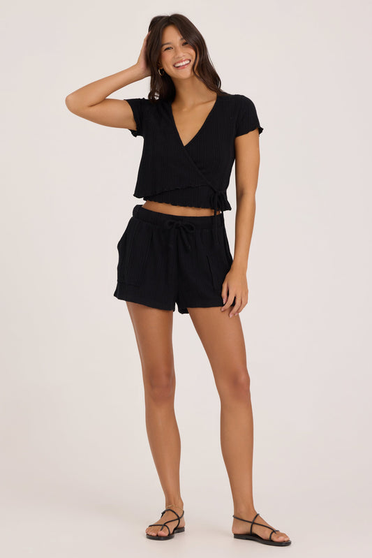 A woman modeling a black two-piece outfit consisting of a short-sleeve wrap-style crop top and matching drawstring shorts, paired with black strappy sandals, against a plain light background.