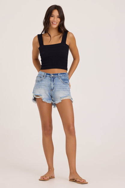 A woman modeling a black ribbed crinkle tank top and light-wash distressed denim shorts against a plain light beige studio background.