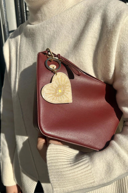 A person in a cream knit sweater holds a structured burgundy leather clutch bag with a gold-tone ring and a beige & gold celestial heart keychain.