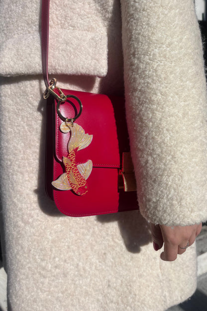Red handbag with a red koi fish keychain held by a person wearing a beige coat.