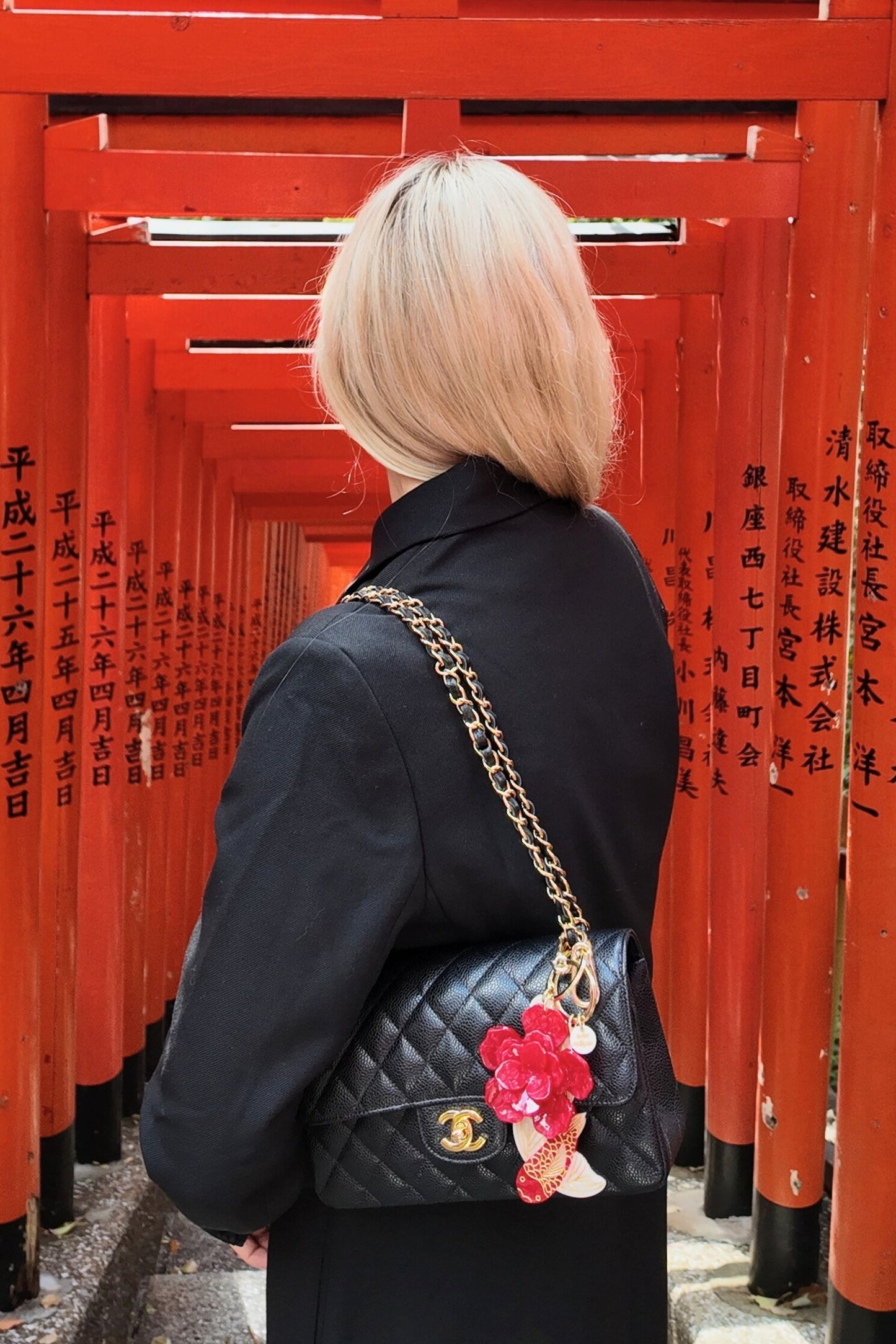 Person with a black quilted handbag featuring a red flower charm and red koi fish keychain  in front of red torii gates.