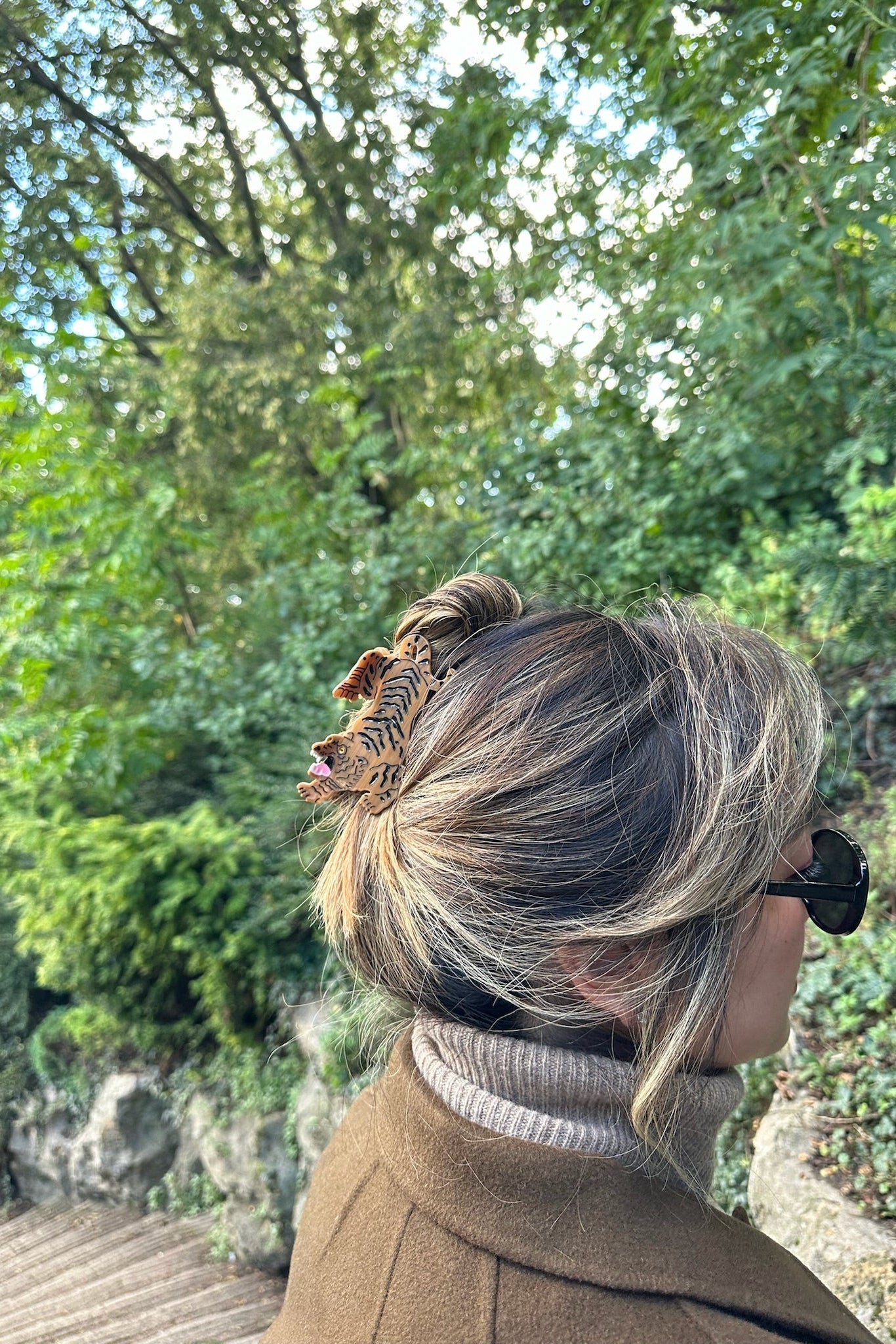 Person with styled hair with a tiger claw clip and sunglasses, standing outdoors with greenery in the background