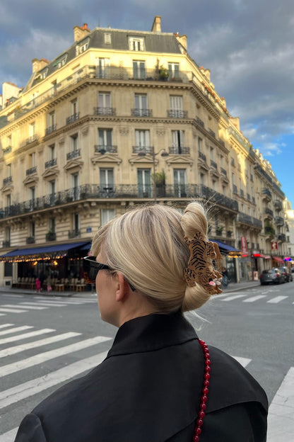 A blonde woman seen from behind wearing a black coat and sunglasses, with her hair styled in a low bun and secured with a tiger hair accessory. She is standing on a city crosswalk in front of a classic Parisian-style building.