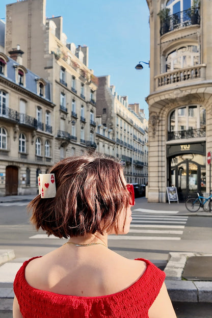 Woman with a red top and Ace of Hearts hair clip standing on a city street with buildings and a crosswalk in the background.