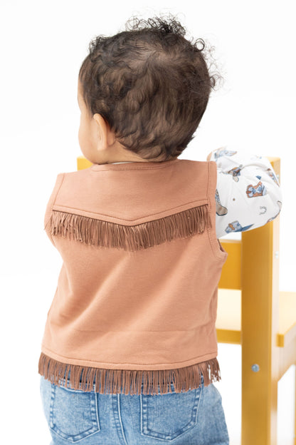 A toddler viewed from behind wearing a solid mocha mousse colored sleeveless vest with fringe trim and blue jeans, standing next to a yellow chair against a white background.