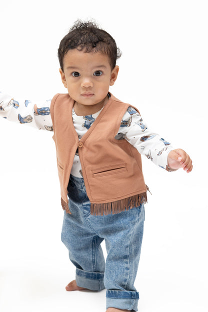 A toddler standing barefoot on a white studio background, wearing a solid mocha mousse colored fringed vest over a white long-sleeve shirt with a blue animal print, blue jeans, and holding onto a yellow chair.