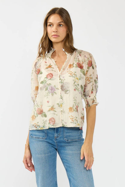 A woman modeling a cream-colored, semi-sheer button-up blouse with a colorful floral print, paired with blue jeans, against a plain light gray background.