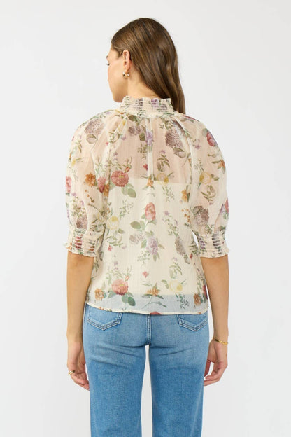 A woman viewed from the back wearing a sheer floral blouse with puff sleeves and a gathered high neck, paired with blue jeans, against a plain light gray studio background.