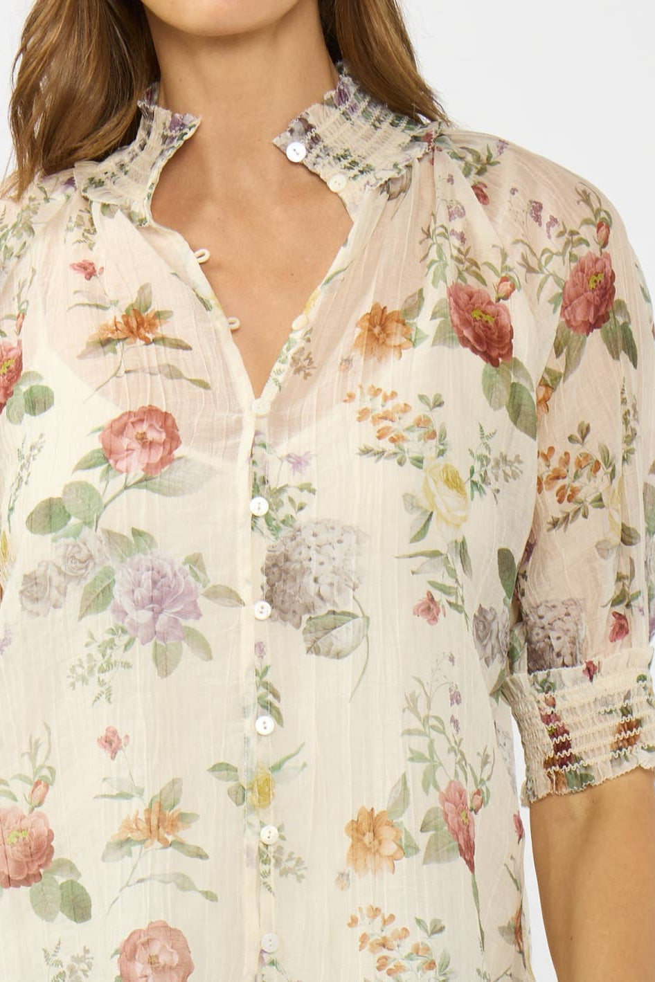 A close-up of a woman wearing a sheer, cream-colored button-up blouse with a delicate pastel floral print and ruffled collar details.