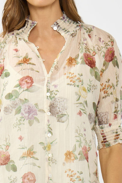 A close-up of a woman wearing a sheer, cream-colored button-up blouse with a delicate pastel floral print and ruffled collar details.