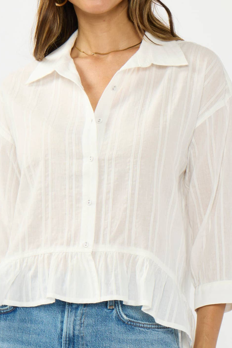 A woman wearing a white semi-sheer long-sleeve button-up blouse with a collar and ruffled hem, paired with blue jeans against a plain white background.