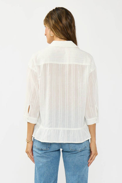 A woman viewed from the back wearing a white sheer long-sleeve blouse with vertical texture details and light blue jeans, set against a plain light gray studio background.