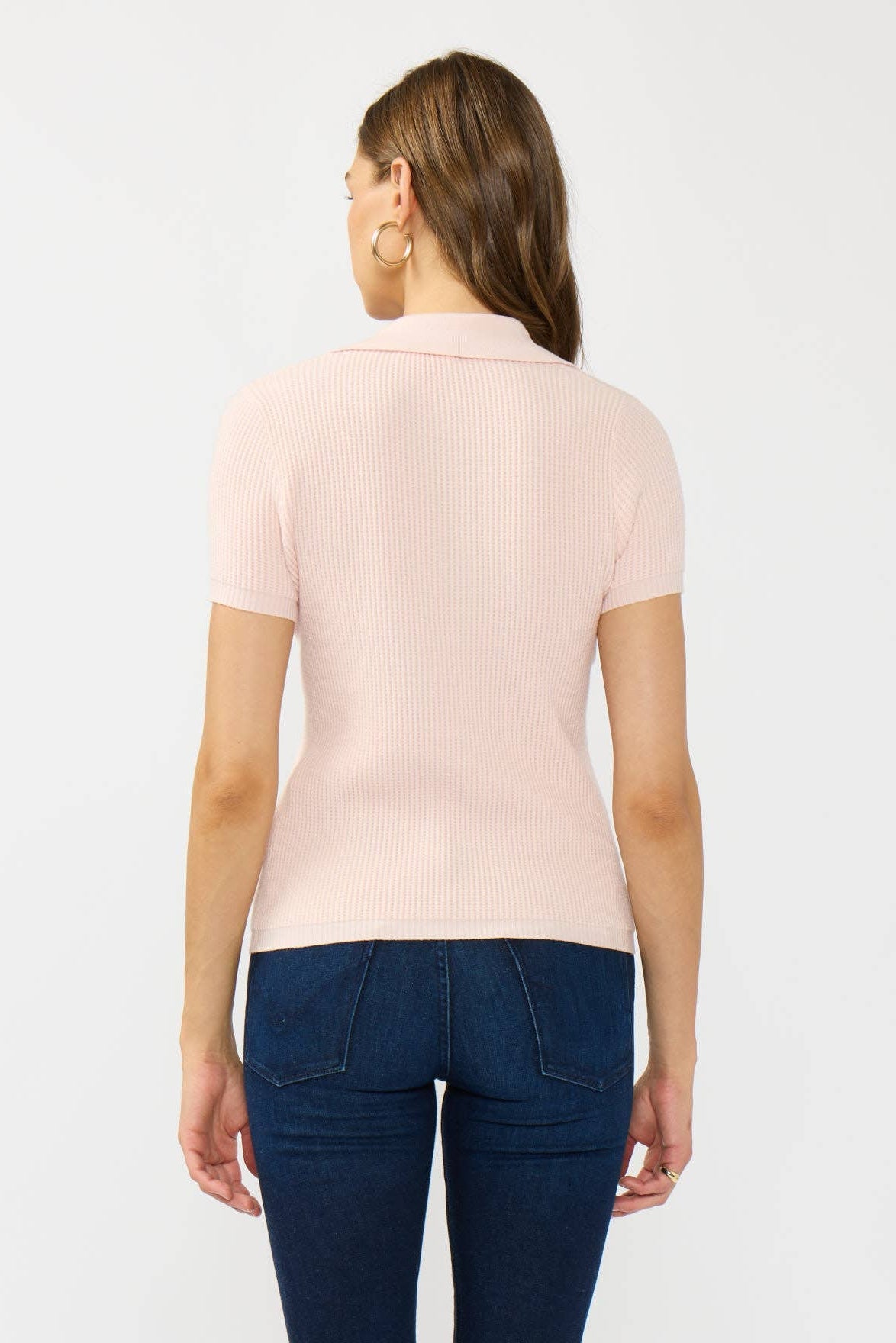 A woman shown from the back wearing a light pink ribbed short-sleeve polo shirt and dark blue jeans against a plain light gray background.