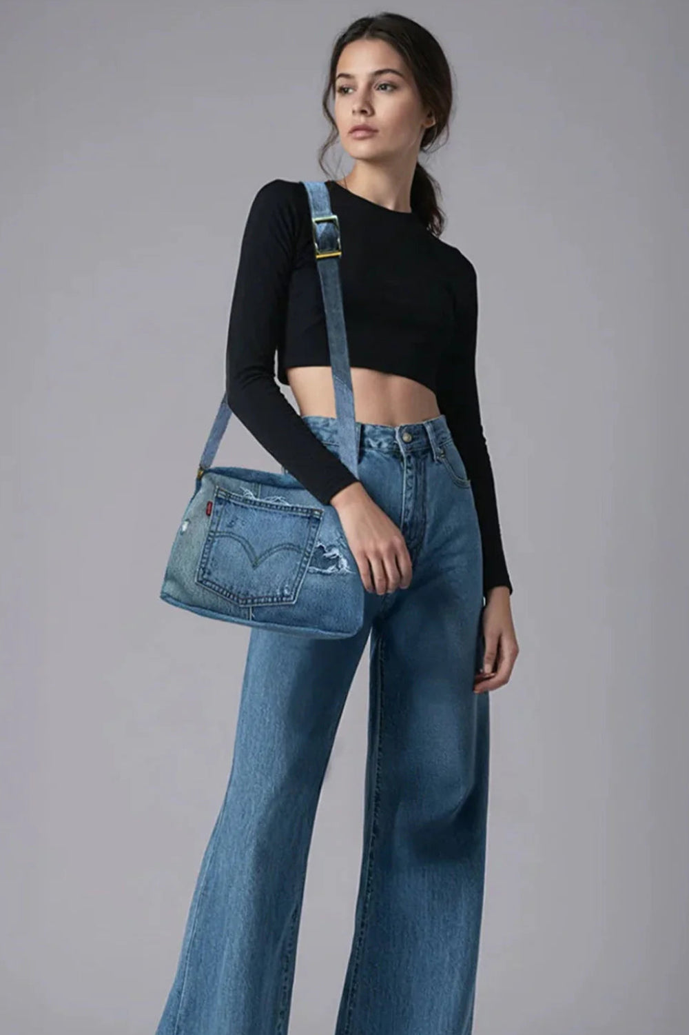 Woman wearing blue jeans and a black top, holding a Conrad denim bag against a gray background