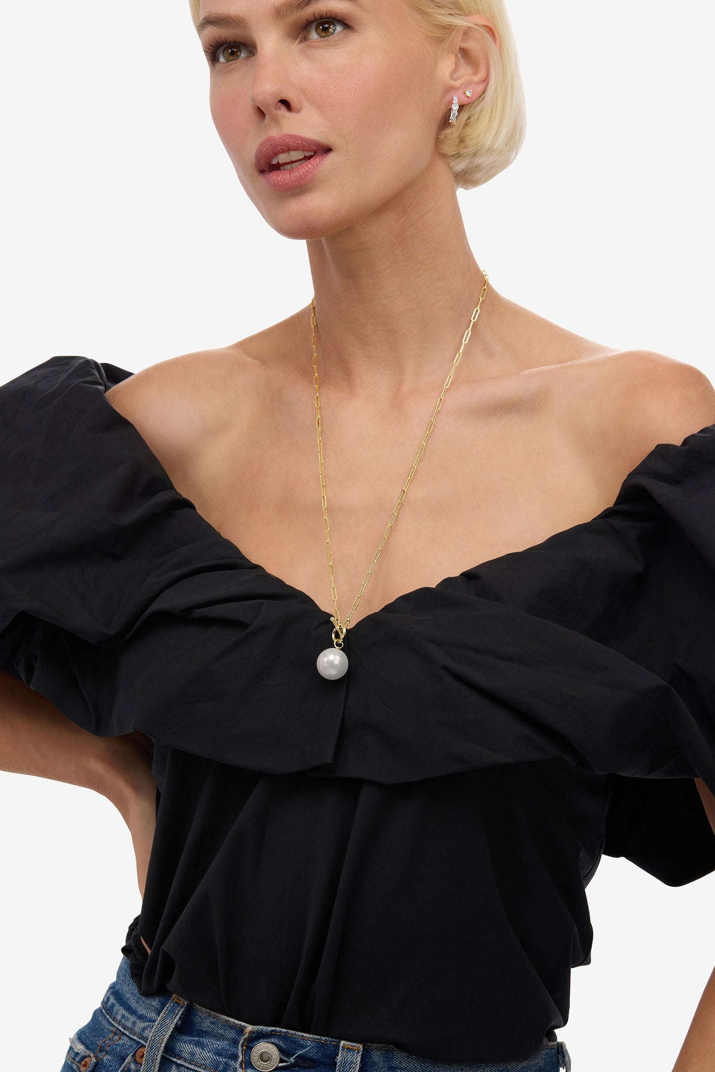 A close-up of a model wearing a black off-the-shoulder blouse with dramatic ruffled sleeves and a gold necklace with a white pearl pendant, paired with blue jeans against a plain white background.