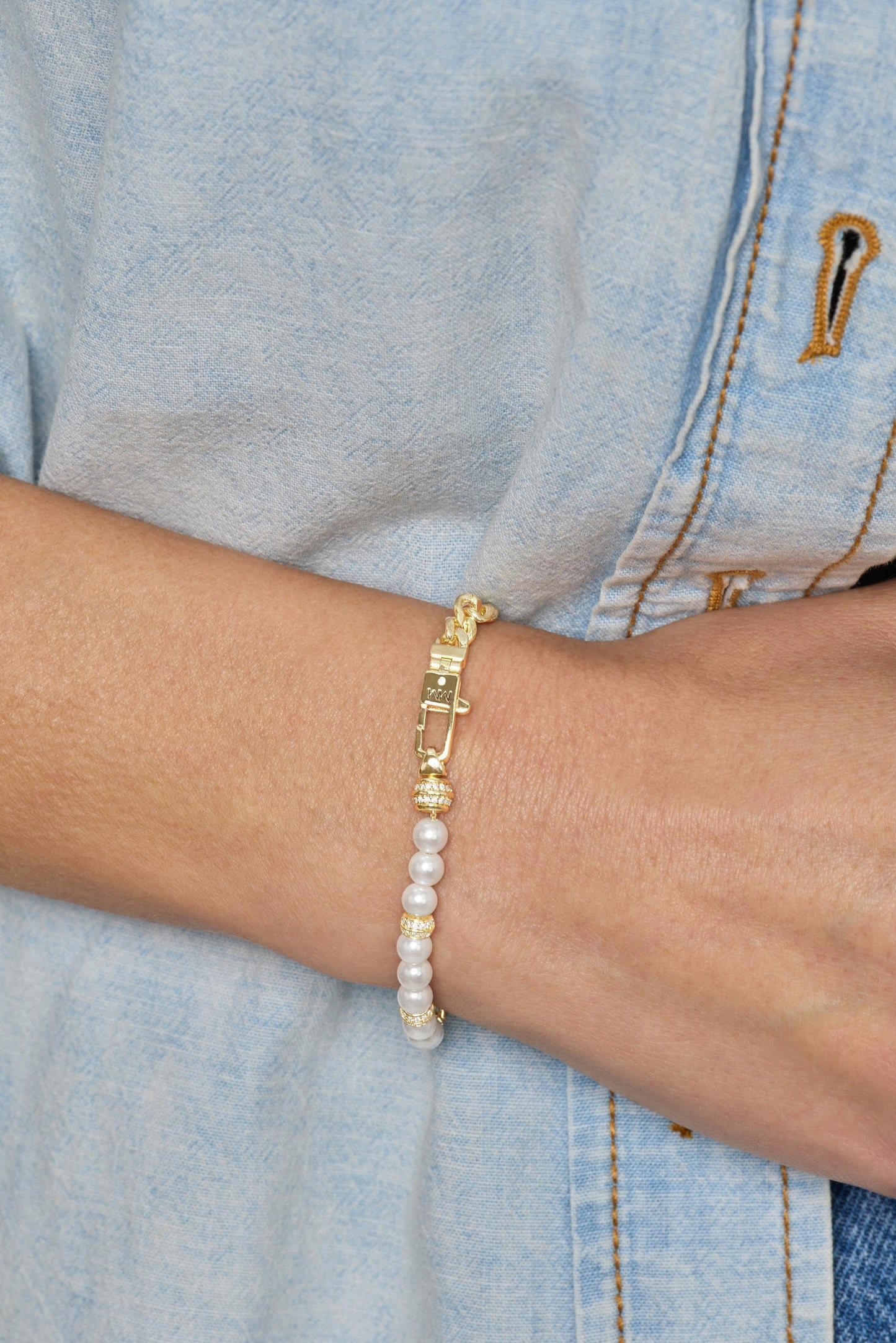 A gold-plated charm bracelet with white pearl-like beads and gold spacer rings, shown worn on a person's wrist over a light blue denim shirt.