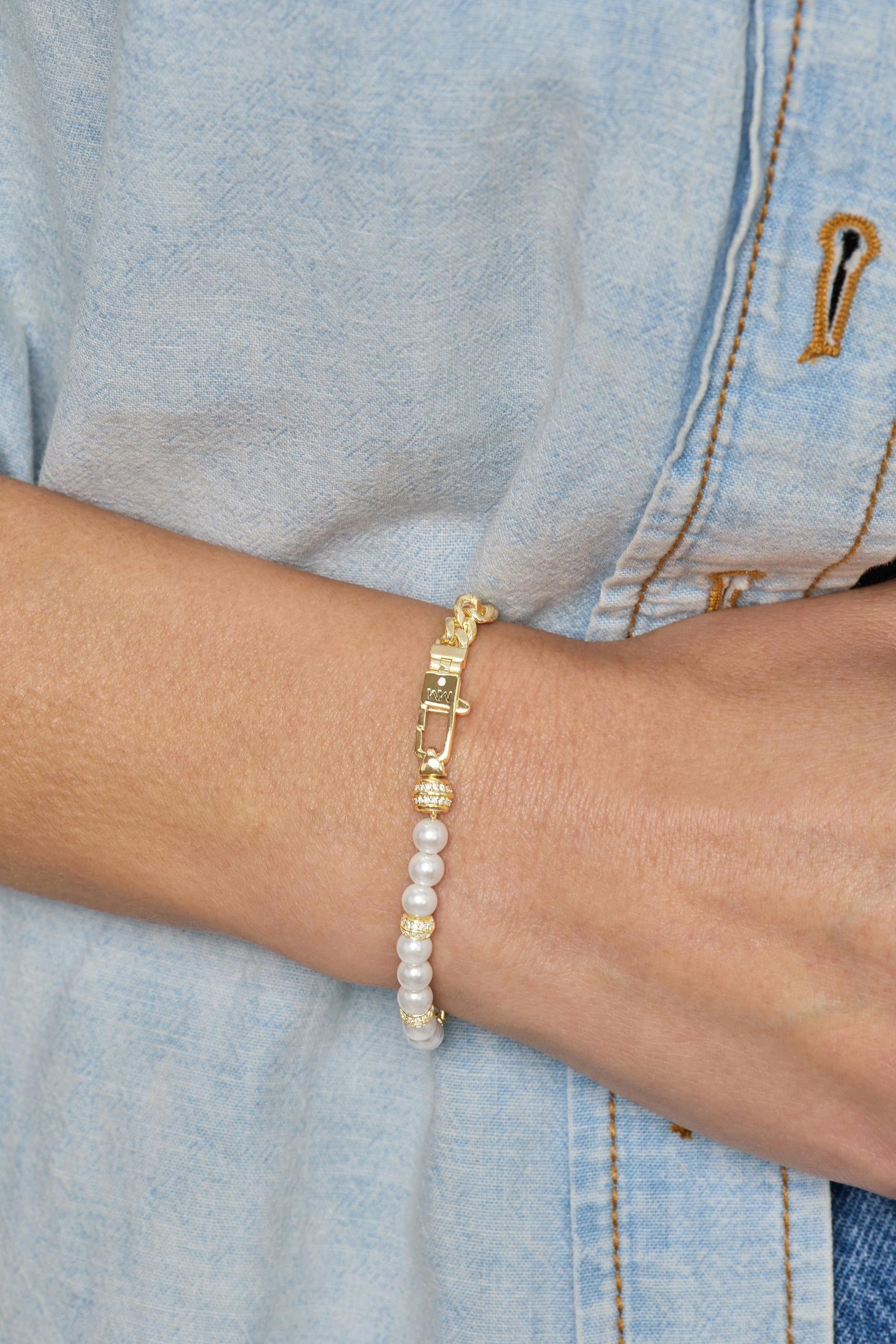 A gold-plated charm bracelet with white pearl-like beads and gold spacer rings, shown worn on a person's wrist over a light blue denim shirt.
