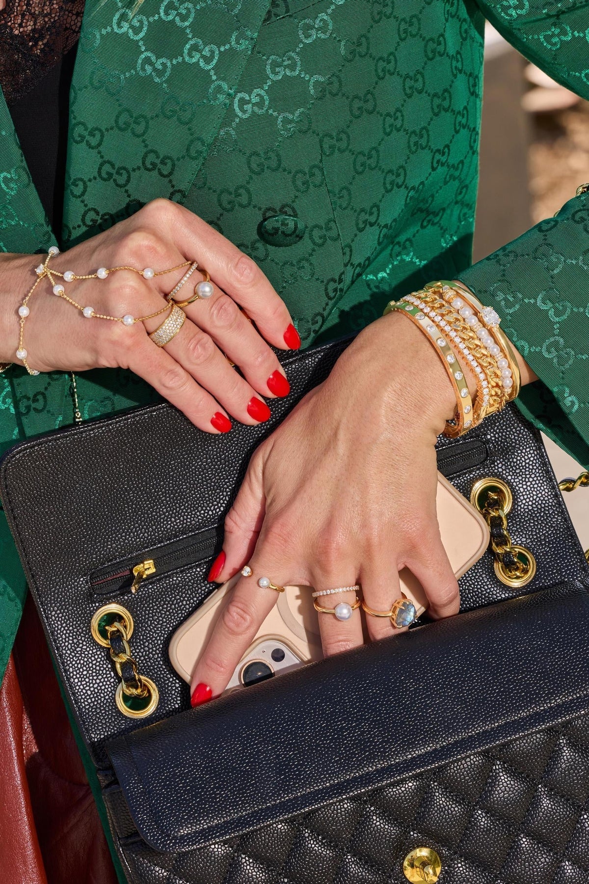 A close-up lifestyle shot of a person wearing a green Gucci jacket and holding a black quilted Chanel shoulder bag with gold hardware. The person's hands are visible, adorned with multiple rings and bracelets, a pearl hand chain and a phone is tucked into the bag's pocket.