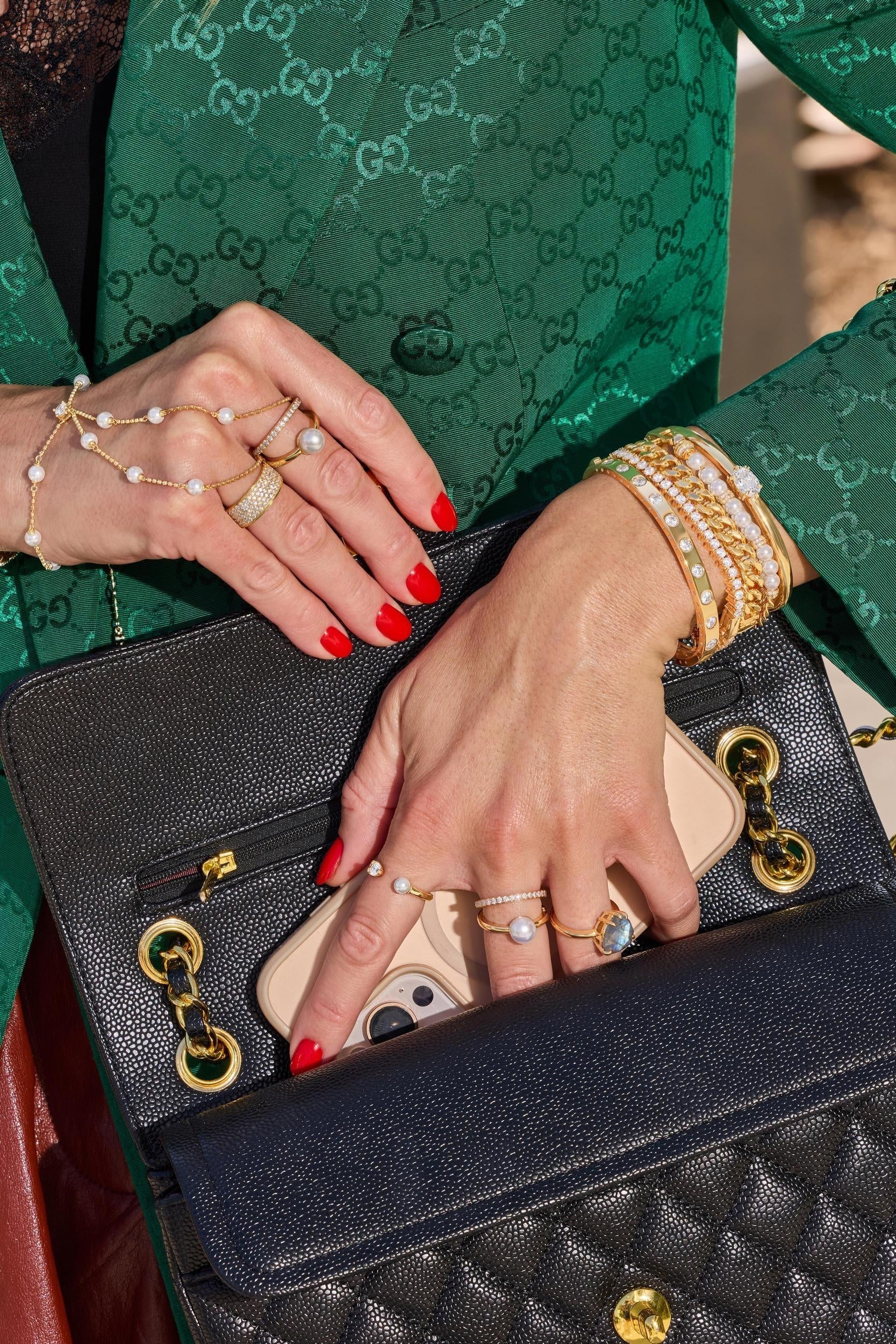 A close-up lifestyle shot of a person wearing a green Gucci jacket and holding a black quilted Chanel shoulder bag with gold hardware. The person's hands are visible, adorned with multiple rings and bracelets, a pearl hand chain and a phone is tucked into the bag's pocket.