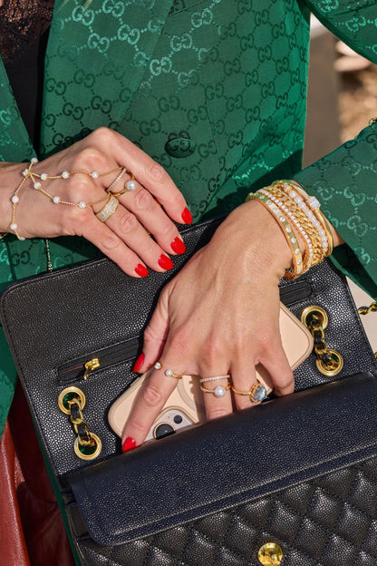A close-up lifestyle shot of a person wearing a green Gucci jacket and holding a black quilted Chanel shoulder bag with gold hardware. The person's hands are visible, adorned with multiple rings and bracelets, a pearl hand chain and a phone is tucked into the bag's pocket.