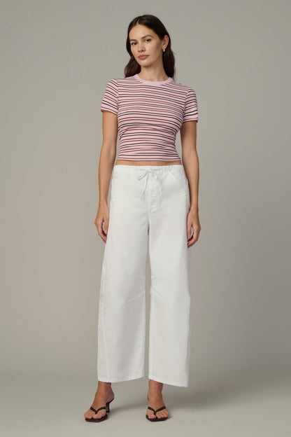 A woman modeling a short-sleeve cropped top with thin blush/ecru horizontal stripes, paired with white wide-leg cropped pants and dark sandals, against a solid gray studio background.
