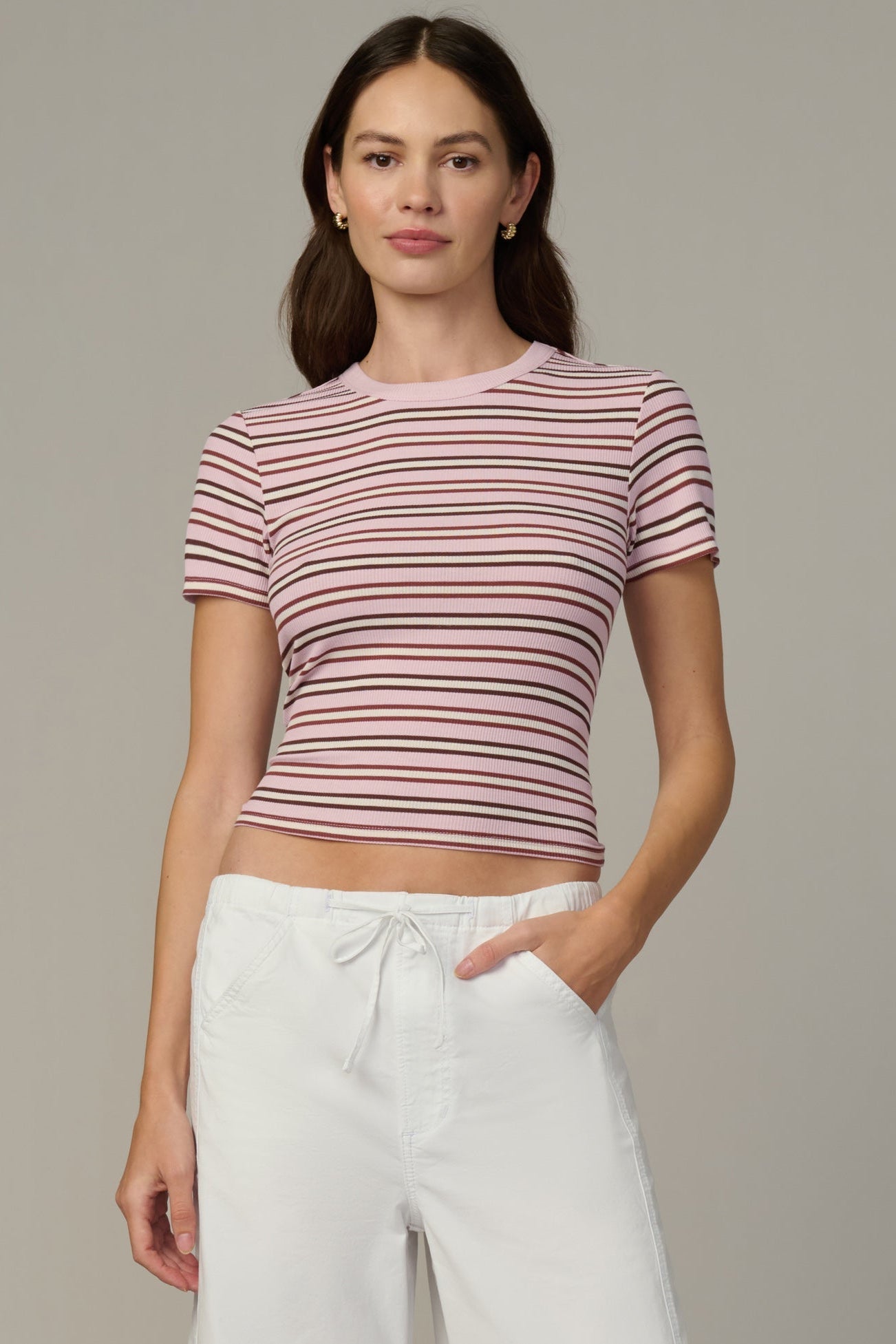 A woman modeling a short-sleeve crewneck crop top with thin horizontal blush/ecru stripes, paired with white drawstring pants, against a solid gray studio background