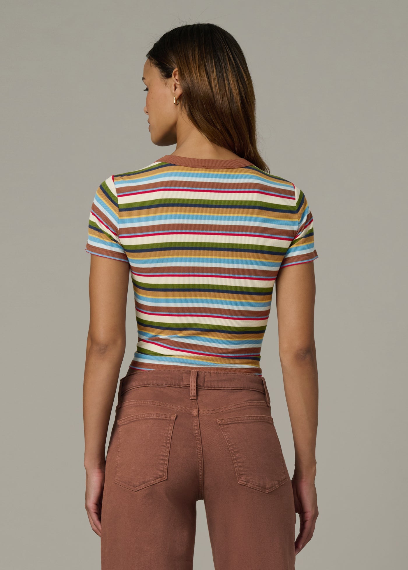Back view of woman wearing a baby tee in multi variegated stripes with brown pants against a neutral background