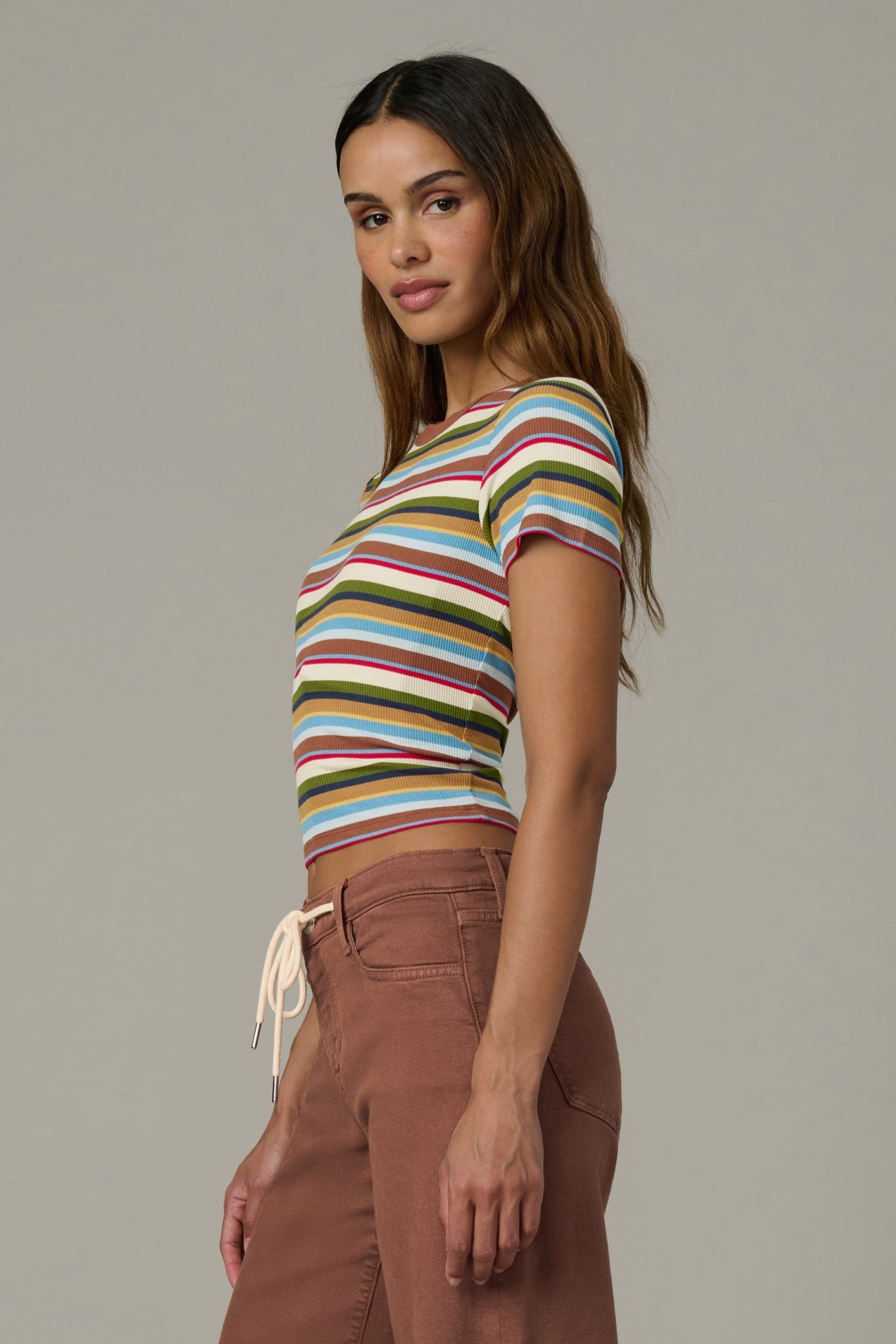 Side view of woman wearing a baby tee in multi variegated stripes with brown pants against a neutral background