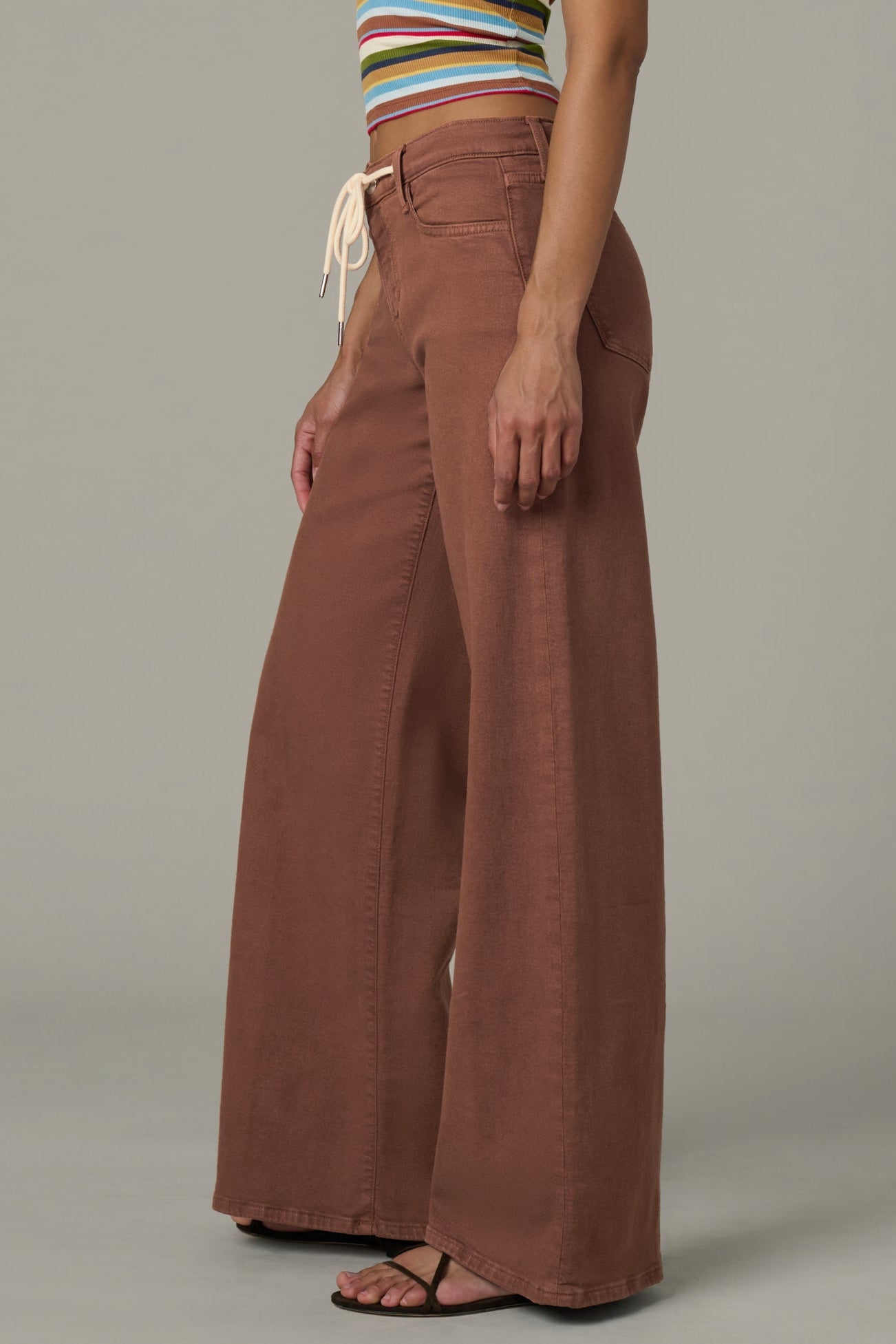 Side view of woman wearing a striped crop top and chocolate color Lou Lou wide-leg pants  with a drawstring on a plain background