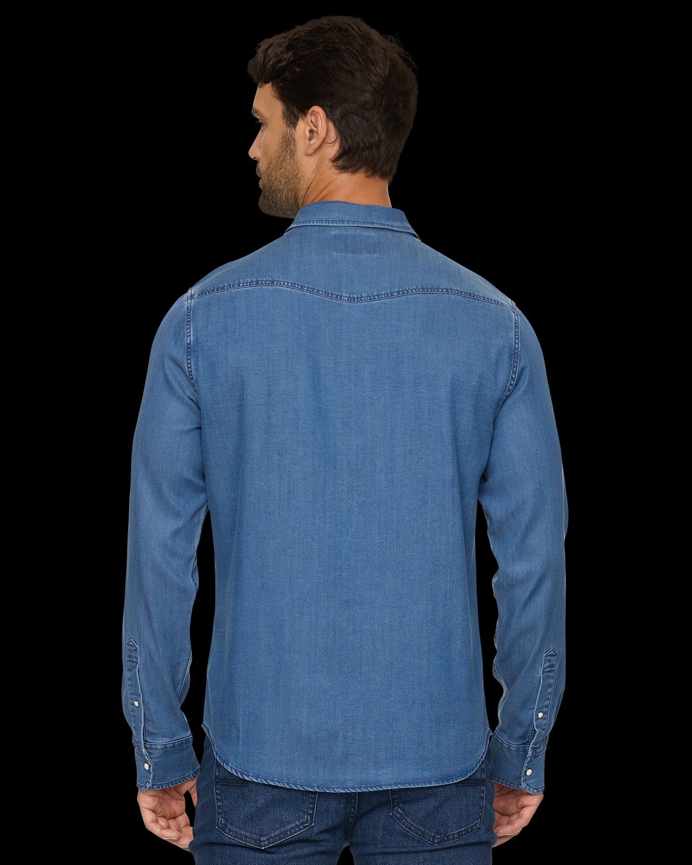A man viewed from the back wearing a medium blue denim long-sleeve button-up shirt and dark blue jeans against a solid black background.