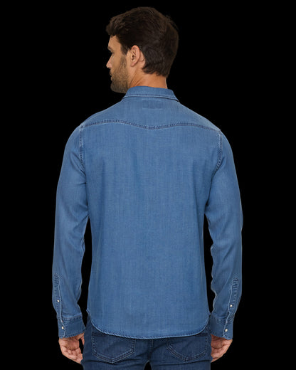 A man viewed from the back wearing a medium blue denim long-sleeve button-up shirt and dark blue jeans against a solid black background.