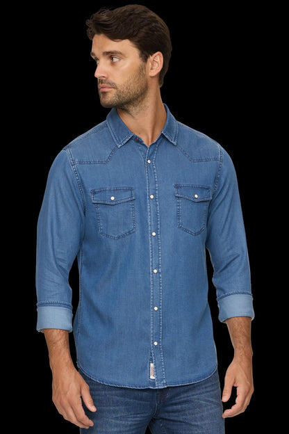 A man wearing a medium-wash blue denim western-style shirt with three-quarter sleeves, rolled cuffs, and two buttoned chest pockets, posed against a solid black background.