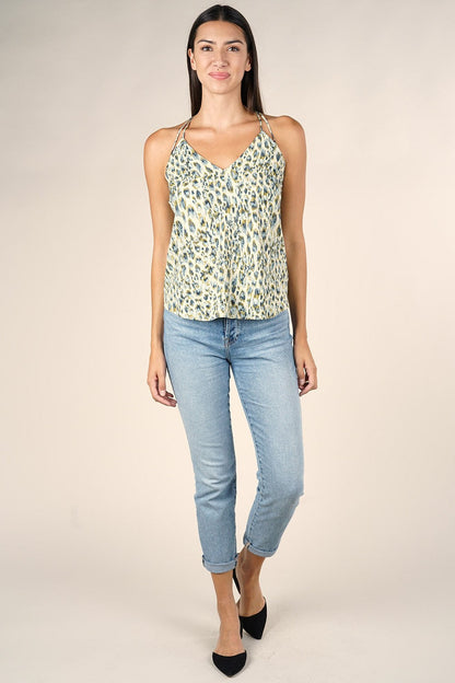 A woman modeling a sleeveless V-neck camisole top with a safari/lagoon pattern, paired with light blue cropped jeans and black heels, against a solid beige studio background.