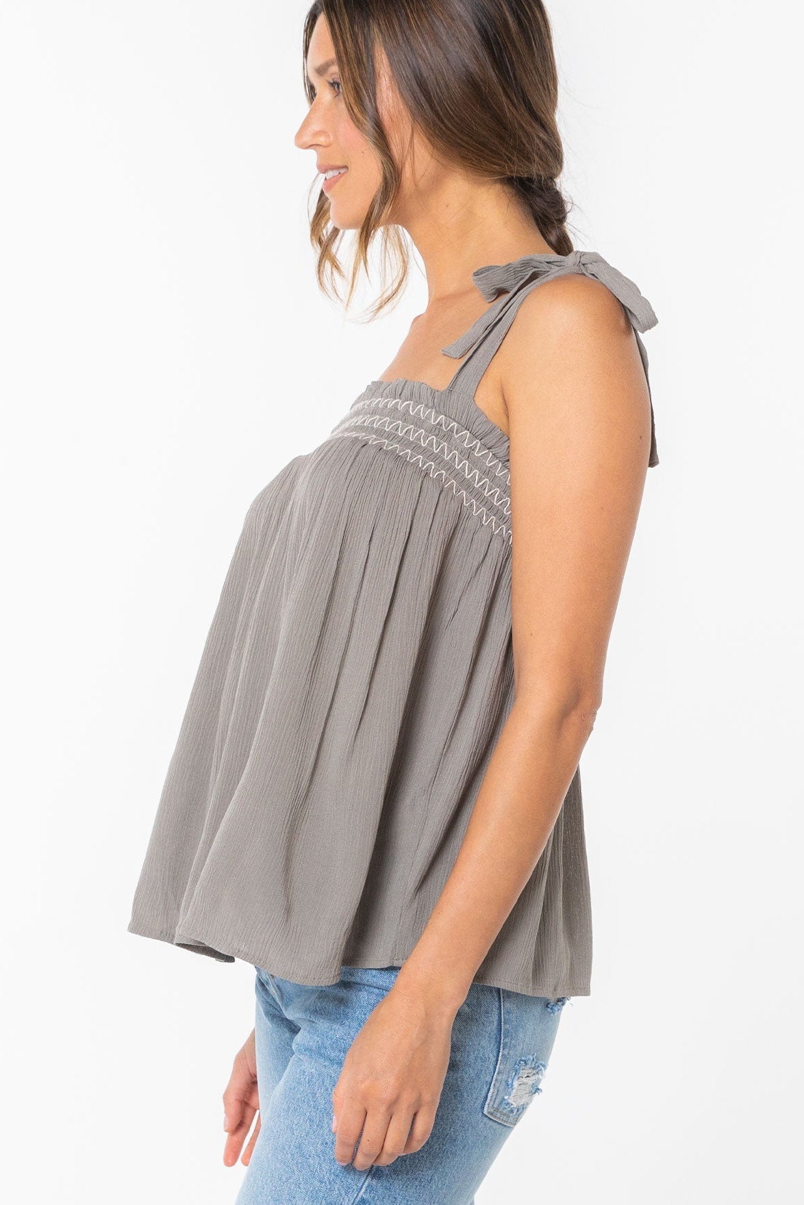 Side view of woman wearing a cement gray sleeveless top with bow details on a white background