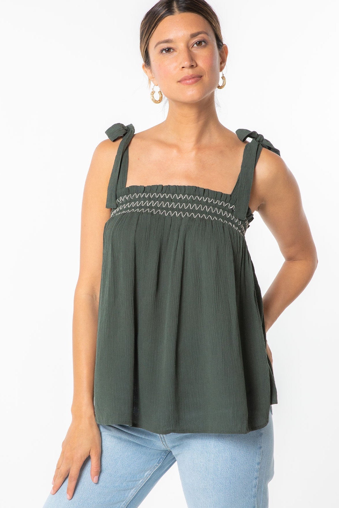 A woman wearing a military green sleeveless top with tie straps and a smocked chest band, paired with light blue jeans, against a plain white background.