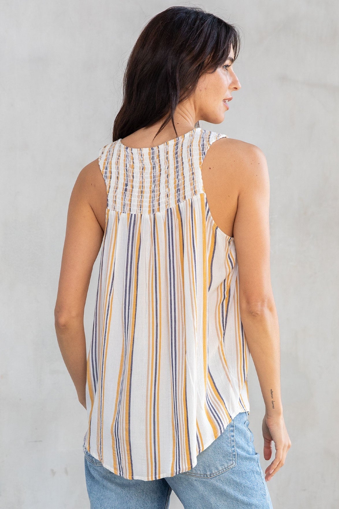 A woman shown from the back wearing a sleeveless top with vertical stripes in cream, dijon yellow, and blue, paired with light blue jeans, against a textured gray wall.