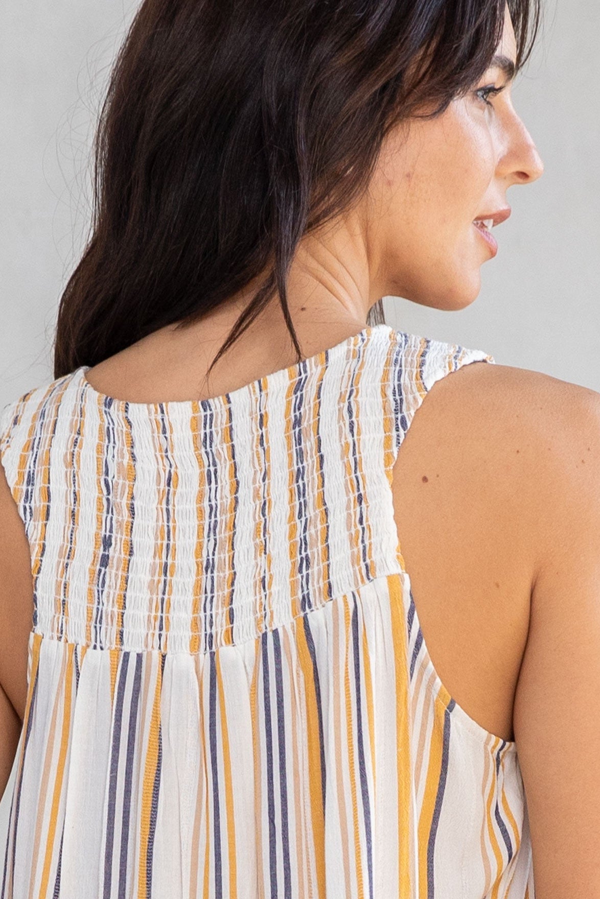 Close-up back view of a woman wearing a sleeveless top with vertical smocked stripes in cream, dijon yellow, and navy blue against a light gray background.