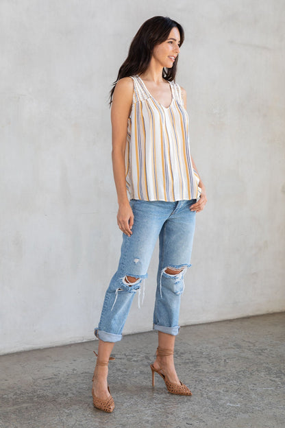 A woman stands in a studio wearing a sleeveless striped top with dijon and blue stripes, distressed cropped jeans and tan high heels against a light gray textured wall.