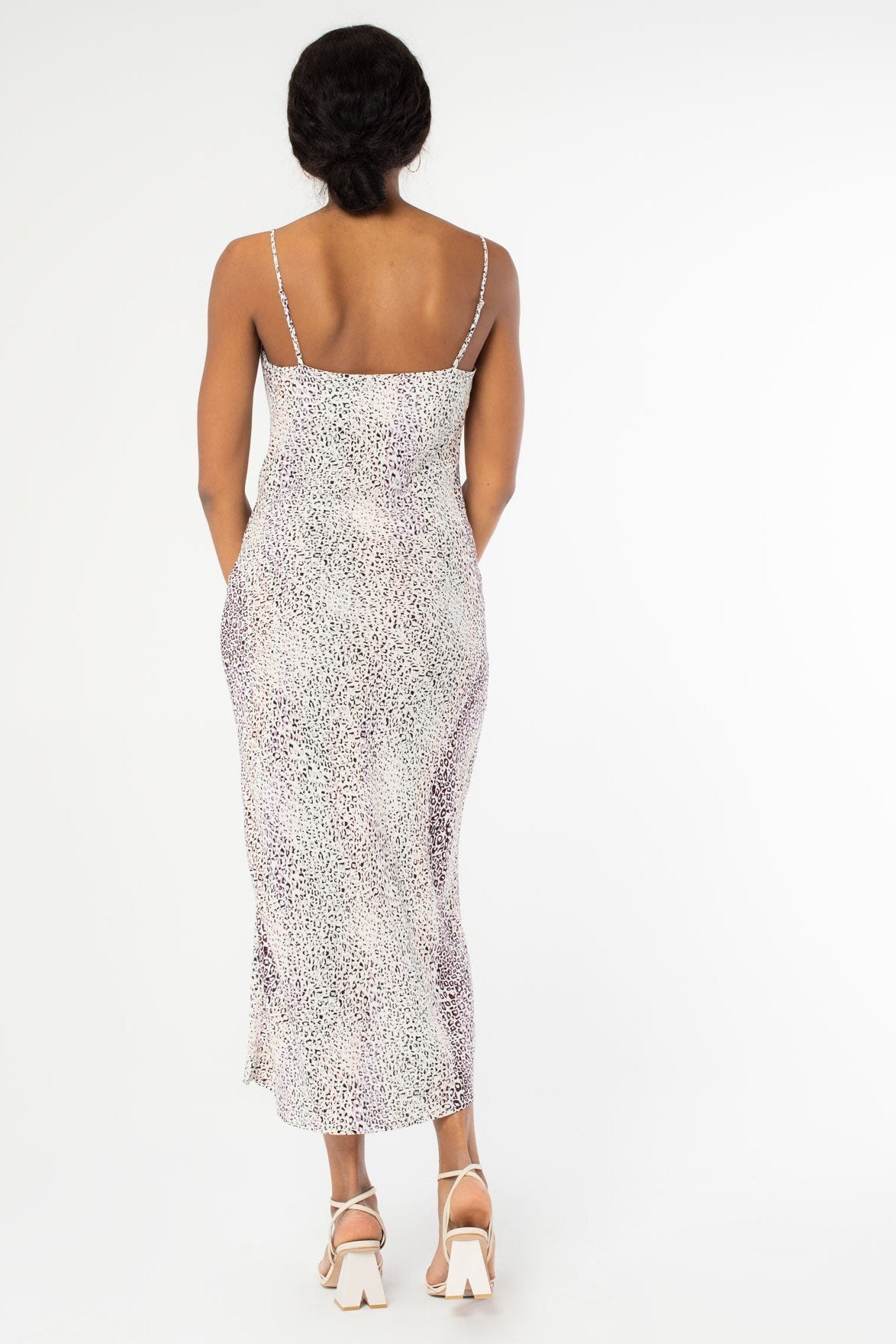 A woman modeling the back of a sleeveless cowl-neck midi slip dress with a lavender and ivory abstract animal pattern, paired with ivory high-heeled sandals, against a plain white studio background.