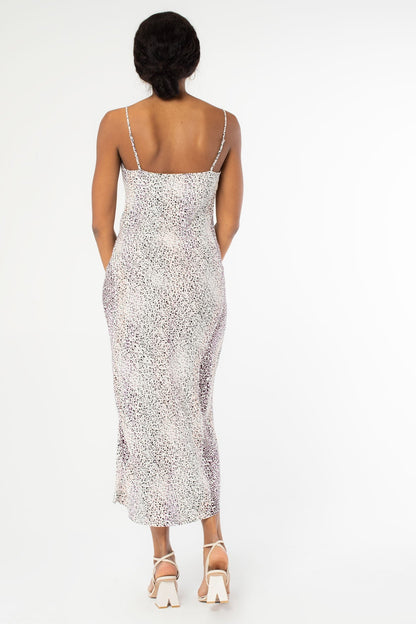 A woman modeling the back of a sleeveless cowl-neck midi slip dress with a lavender and ivory abstract animal pattern, paired with ivory high-heeled sandals, against a plain white studio background.