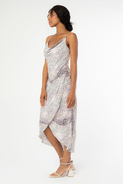 Side view of a woman modeling a sleeveless cowl-neck midi slip dress with a lavender and ivory abstract animal pattern, paired with ivory high-heeled sandals, against a plain white studio background.